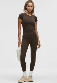 Naine pruunis sportlikus riietuses hoiab heleda veepudelit, tal on jalas valged sokid ja heledad tossud, seismine ühetaolise tausta ees.