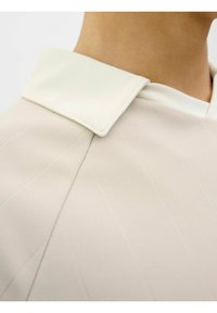 Lys beige skjorte med en kontrasterende hvid krave, der har struktureret stof og diagonale sømdetaljer for et struktureret udseende.