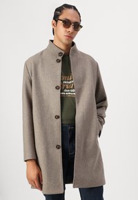Brun ullfrakk med ståkrage, enkel knappestengning og store knapper. Baird over en grønn T-skjorte og mørke jeans.