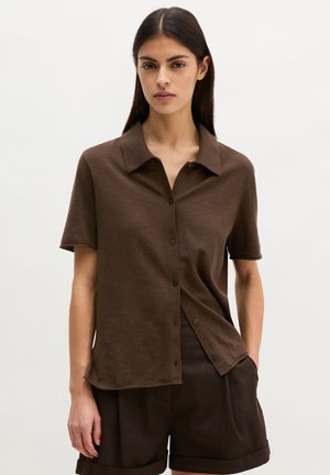 Vrouw die een bruin kortermig overhemd met knopen en bijpassende bruine korte broek draagt, staand tegen een effen lichte achtergrond.