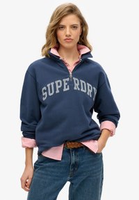 Granatowa bluza zapinana na zamek z napisem "SUPERDRY" w jasnoszarym kolorze, zestawiona z jasnoróżową koszulą z kołnierzykiem i dżinsami.