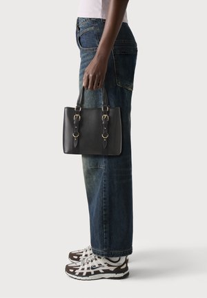 Black leather handbag with two gold-tone buckles, rectangular shape, and short handles, held by a person in denim jeans and sneakers.