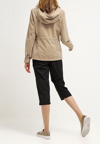 Giacca a vento beige chiaro con cappuccio, vita elastica, maniche lunghe e polsini con bottone, abbinata a pantaloni neri a tre quarti e scarpe gold slip-on.