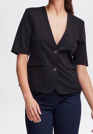 Femme portant un blazer noir à manches courtes avec deux boutons et un pantalon bleu marine, debout devant un fond clair uni.