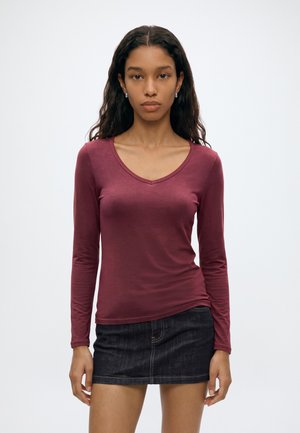 Jeune femme aux longs cheveux foncés portant un haut bordeaux à manches longues avec un col en V et une mini-jupe en denim foncé sur un fond uni.
