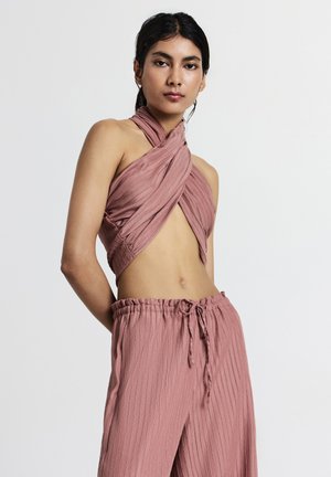 Donna che indossa un top corto a halter plissettato color rosa antico abbinato a pantaloni larghi con coulisse su uno sfondo bianco semplice.