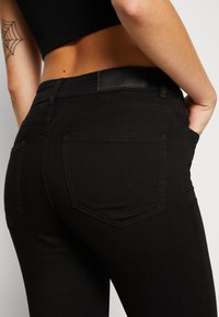 Jeans de mezclilla negra con una textura suave, corte de altura media y dos bolsillos traseros. Presenta costuras sutiles y un parche de cuero en la cintura.