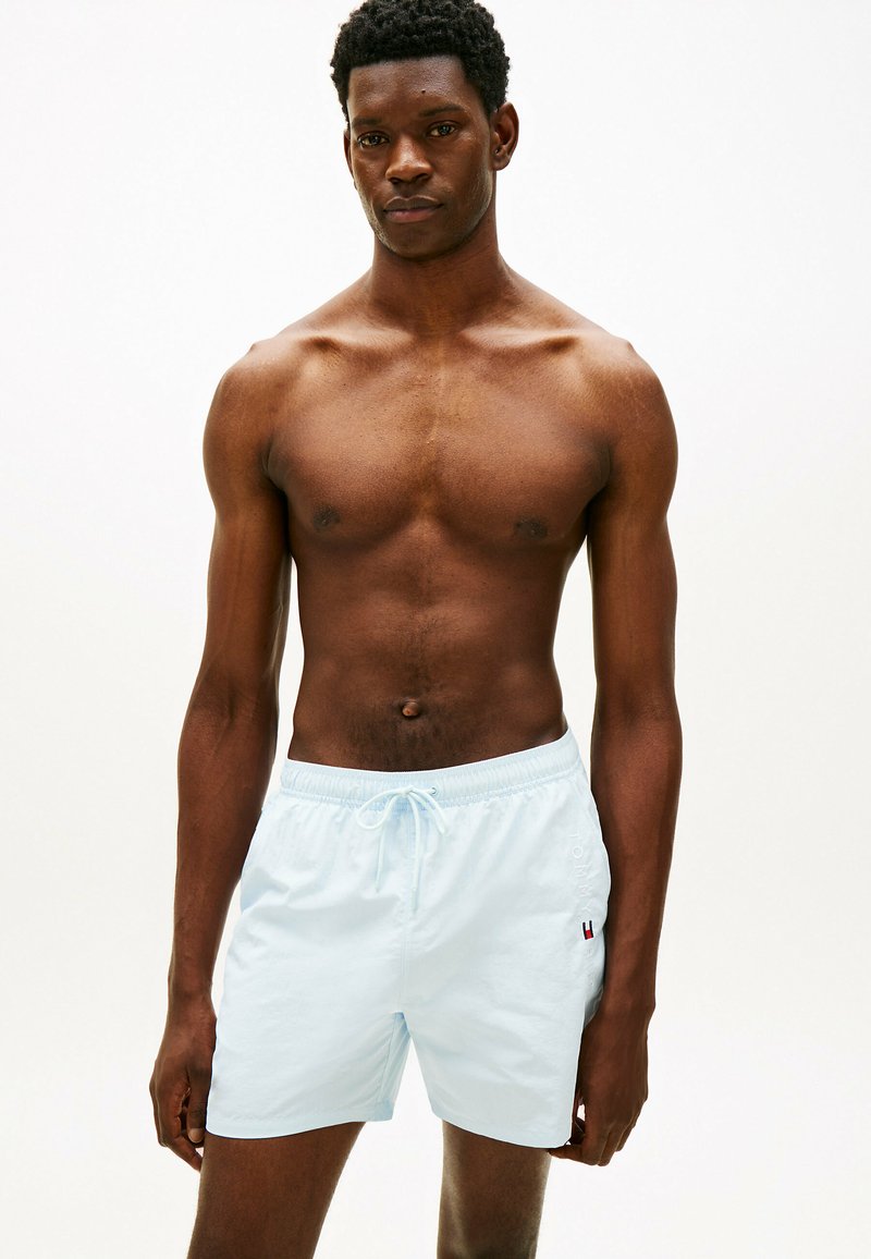 Homme musclé debout, bras détendus, portant un short de bain blanc à cordon, sur un fond clair uni.