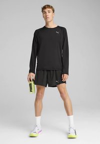 Sort langærmet skjorte og shorts, begge med en reflekterende logo. Hvide sokker og sko med gule detaljer og logo. Gennemsigtig vandflaske i hånden.