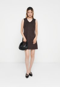 Brown sleeveless dress with a V-neck design. Model holds a black leather handbag. Footwear features pointed toes and shiny texture.