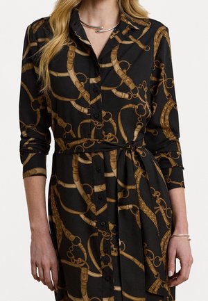 Femme portant une robe noire boutonnée à manches longues avec une ceinture équestre marron et un imprimé chaîne, nouée à la taille avec une ceinture en tissu assorti.