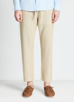 Mann trägt beige, gerade geschnittene Hose und braune Leder-Schnürschuhe, steht vor neutralem Hintergrund.