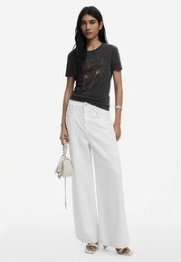 Grafisk t-shirt i mörkgrå med palmmönster i metallicguld. Kombinerad med vida vita byxor och en liten vit handväska.