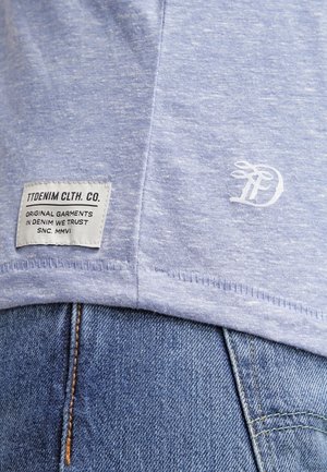 Personne portant un t-shirt bleu clair avec une étiquette en tissu et un logo blanc sur un jean bleu, mettant l'accent sur l'ourlet du t-shirt et la ceinture du jean.