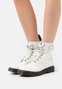 Białe skórzane botki na sznurowadła ze srebrnymi elementami, wyściełanym kołnierzem i czarną podeszwą z żółtym przeszyciem.