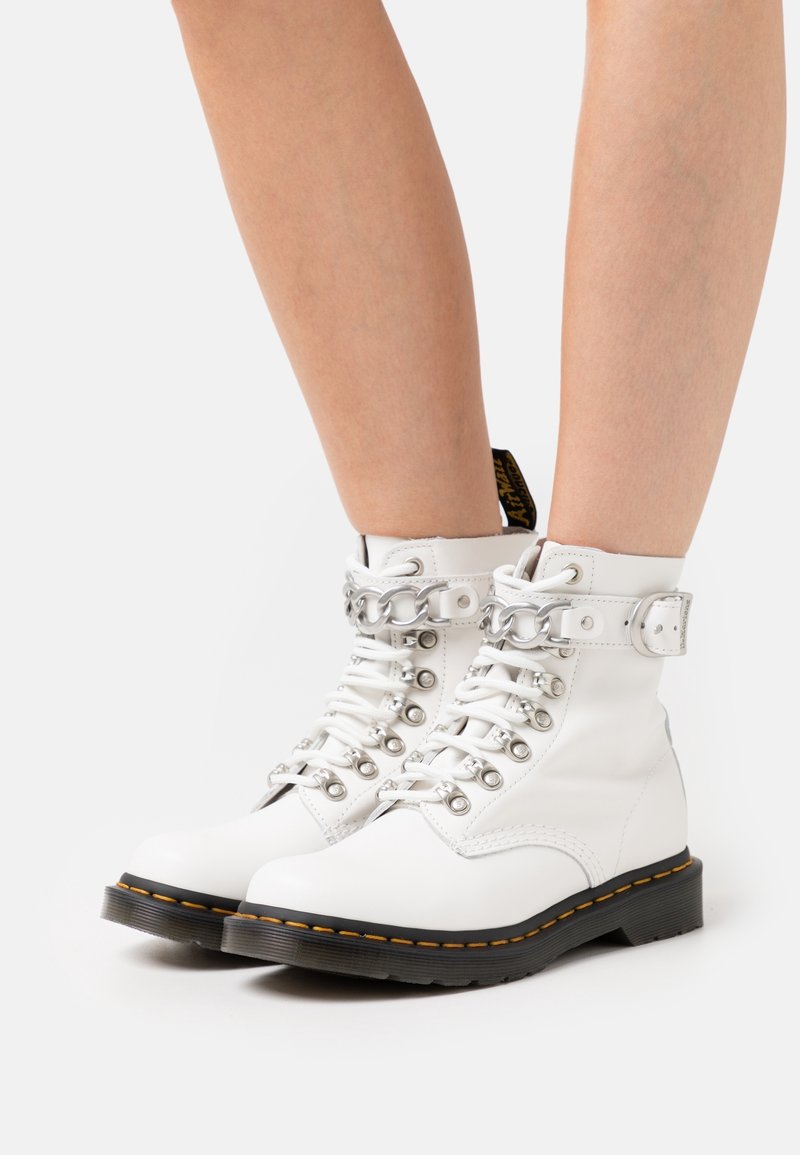 Białe skórzane botki na sznurowadła ze srebrnymi elementami, wyściełanym kołnierzem i czarną podeszwą z żółtym przeszyciem.