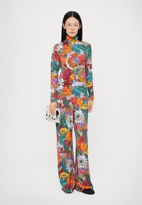 Top cu imprimeu floral, cu mâneci lungi și pantaloni largi în culori vibrante. Modelul ține o poșetă cu model. Material neted, design ajustat, aspect modern.