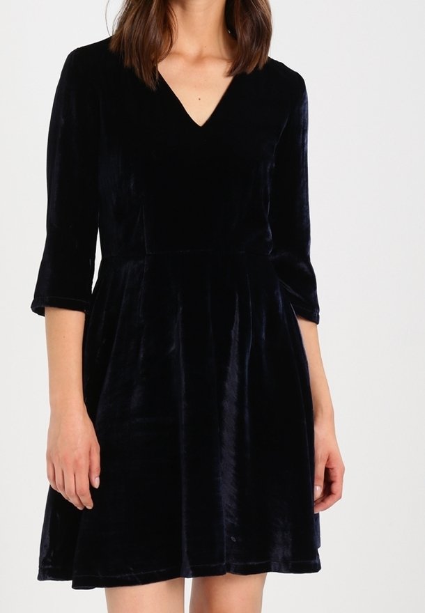 Mujer vistiendo un vestido de terciopelo negro hasta la rodilla con mangas tres cuartos y un escote en V frente a un fondo blanco liso.