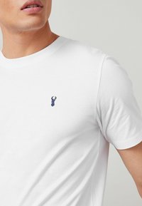 Camiseta blanca de algodón con un pequeño bordado de langosta azul en el pecho. Cuello redondo clásico y manga corta. Textura suave.
