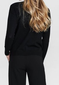 Femme blonde portant un pull à manches longues noir texturé et un pantalon noir, debout les mains dans les poches, dos à la caméra.