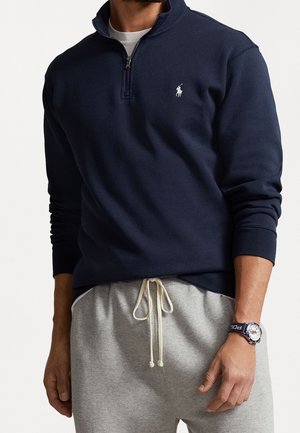 Mężczyzna w granatowej bluzie na zamek z białym logo, jasnoszarych spodniach dresowych ze sznurkiem oraz zegarku na nadgarstek z granatowym paskiem "Polo".