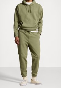 Olivově zelená mikina s kapucí a odpovídající jogger kalhoty vyrobené z měkké tkaniny. Obsahuje přední kapsu, šňůrku v pase a elastické man žety. Součástí jsou také pohodlné boty.