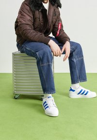 Osoba v hnědém kabátu s kožešinovým límcem, modrých džínách a bílých teniskách Adidas sedí na kovovém kufru s kolečky na zelené podlaze.