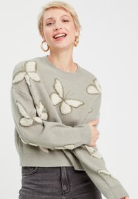 Pull gris avec des motifs de papillons crémeux texturés, col rond et manches longues. Le matériau semble doux et confortable. Coupe courte.