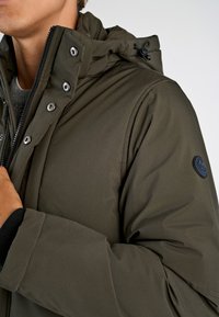 Olivenfarvet isoleret jakke med lynlås og trykknaplukning, med hætte, sølvfarvet hardware og et rundt mærke på ærmet.