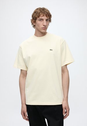 Młody mężczyzna o jasnych kręconych włosach, ubrany w jednolity jasnobeżowy t-shirt z krótkim rękawem i czarne spodnie, stojący na białym tle.