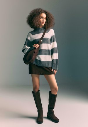 Woman with curly hair wearing a gray and white striped sweater, dark skirt, knee-high brown boots, and carrying a brown shoulder bag.