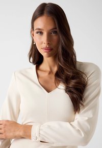 Młoda kobieta o długich brązowych włosach nosi perłowe kolczyki i kremową bluzkę z dekoltem w serek oraz ozdobnymi guzikami na mankietach, na jednolitym tle.