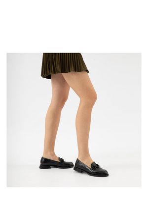 Nogi w czarnych skórzanych mokasynach z ozdobnym łańcuszkiem i spódnicy z oliwkowej zieleni plisowanej, stojące na białym tle.