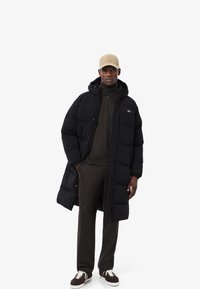 Abrigo acolchado negro con capucha, con secciones acolchadas y un pequeño logo. Usado sobre un suéter oscuro y pantalones a juego, combinado con zapatos marrones.