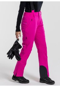 Heldere roze skibroek met verstelbare bretels, zijritszakken, versterkte onderbenen, en gecombineerd met zwarte handschoenen en stevige zwarte laarzen.