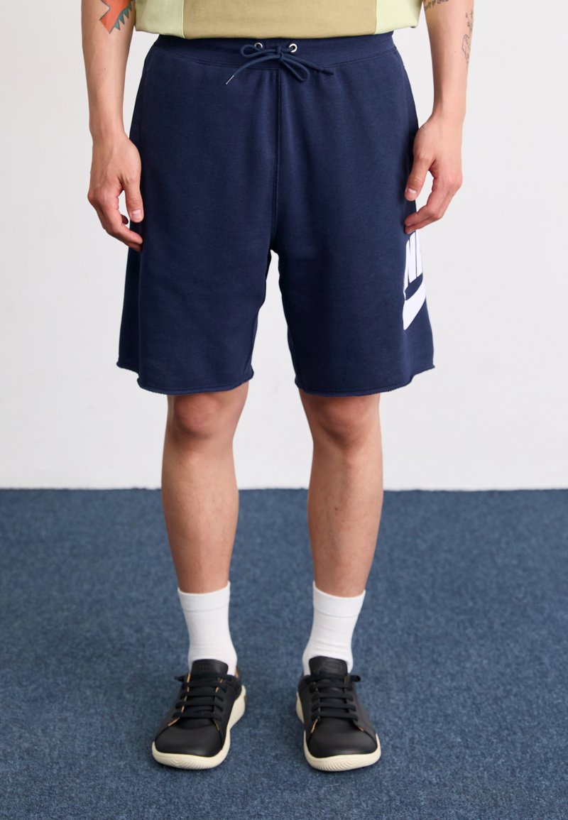 Námornícke modré šortky Nike, vyrobené z jemného materiálu, majú pás s nastaviteľným šnúrkom a biele logo ako akcent. Nosené s čiernymi teniskami a bielymi ponožkami.