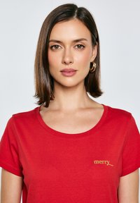 Červené bavlněné tričko s krátkým rukávem, kulatým výstřihem a zlatě vyšitým textem "merry" na levé straně, hladká textura.