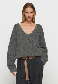 Grauer, flauschiger Strickpullover mit tiefem V-Ausschnitt und langen Ärmeln, kombiniert mit hochgeschnittenen, dunklen Hosen, die einen kontrastierenden Gürtel besitzen.