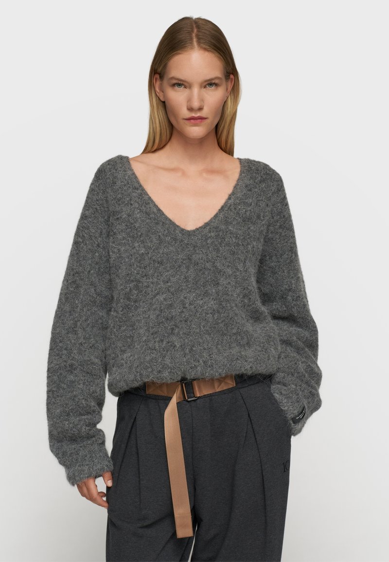 Grauer, flauschiger Strickpullover mit tiefem V-Ausschnitt und langen Ärmeln, kombiniert mit hochgeschnittenen, dunklen Hosen, die einen kontrastierenden Gürtel besitzen.