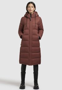 Brūns garš pūkains mētelis ar pieguļošu kapuci, kam ir rāvējslēdzēja aizdare, liels horizontāls quiltēšanas raksts, divas sānu kabatas un regulējamas saites.