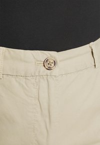 Bežové kalhoty s hladkou strukturou, vybavené zapínáním na knoflík a prošitým pasem. Tkanina má mírně pomačkaný vzhled.