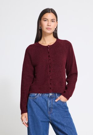 Femme aux longs cheveux foncés portant un cardigan bordeaux boutonné et un jean bleu, debout, une main dans la poche, sur un fond uni.