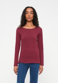 Bordeaux langærmet top i en tætsiddende design, lavet af blødt bomuldsstof, med rund halsudskæring og lige kant. Kombinationen med blå denim er synlig.