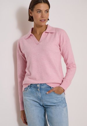 Kvinde iført en lyserød langærmet V-hals sweater og lyseblå jeans, stående med den ene hånd i lommen foran en ensartet baggrund.