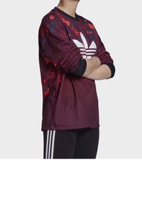 Persona che indossa una maglietta a maniche lunghe Adidas color bordeaux con logo trefoil bianco e pantaloni neri con strisce bianche, braccia incrociate.
