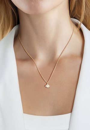 Delikat guldkæde med en lille, vifteformet vedhæng, båret om en kvindes hals over en hvid blazer og top.