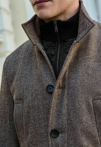 Ruskea herringbone-kuvioinen takki, jossa on korkea kaulus, musta vetoketjullinen sisätakki, kaksi nappia ja sivutasku.