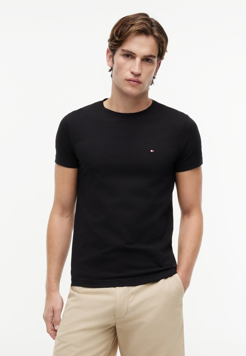 Junger Mann trägt ein schwarzes kurzärmeliges T-Shirt mit kleinem Logo auf der Brust und beigen Hosen, steht vor einem schlichten weißen Hintergrund.