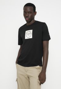 Μαύρο βαμβακερό μπλουζάκι με ένα εκρού τετράγωνο και μαύρο κείμενο. Κοντά μανίκια, στρογγυλή λαιμόκοψη και μινιμαλιστικός σχεδιασμός. Άνετη εφαρμογή.