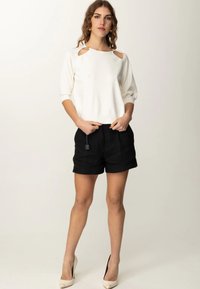 Top bianco con aperture sulle spalle e dettagli dorati, abbinato a pantaloni corti neri. Modella che indossa scarpe con tacco crema, ambientata su uno sfondo neutro.